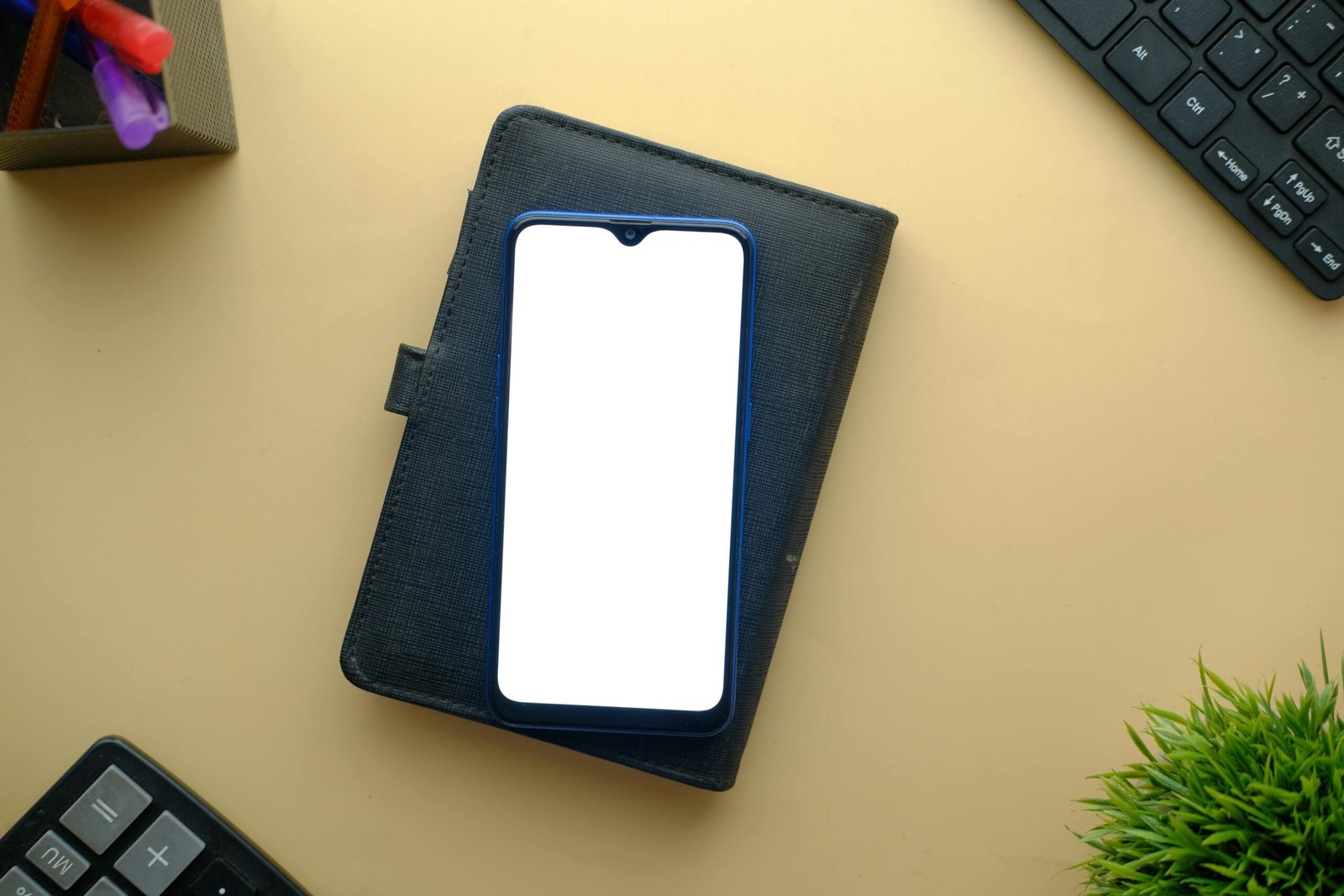 Flat lay of smartphone with blank screen on styled desk with keyboard and plant. Perfect for mockup displays.