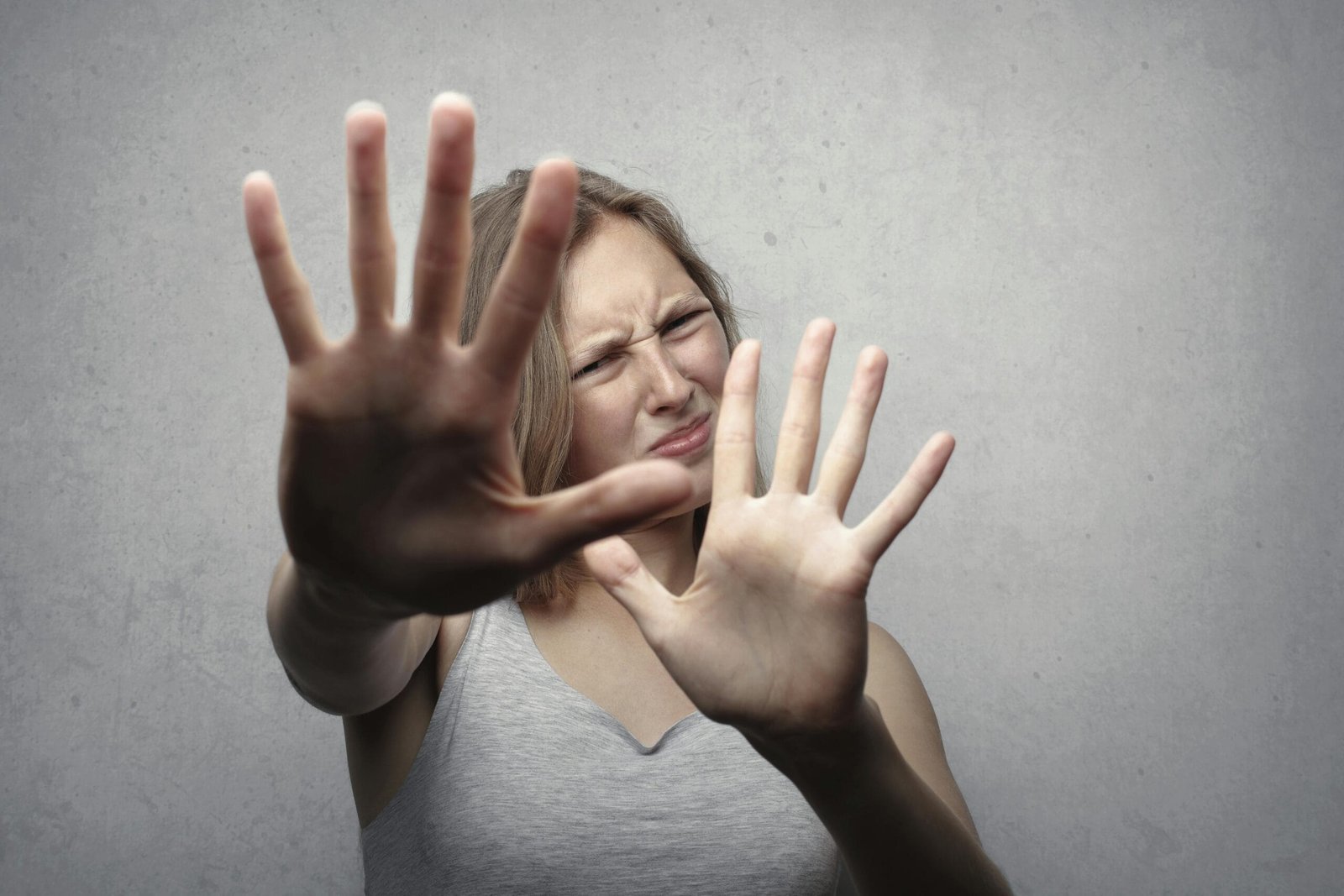 Portrait of a fearful woman in a gray tank top with hands pushed forward against a gray background.