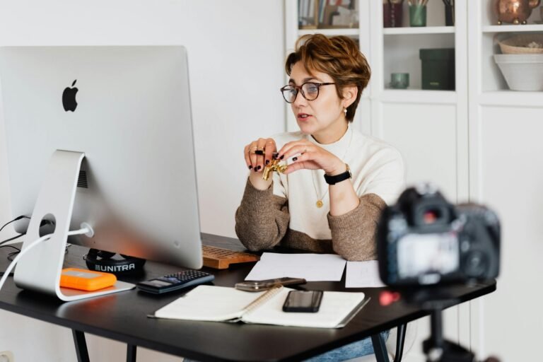 A professional woman engaged in a virtual meeting setup at her home desk, using a computer and camera.