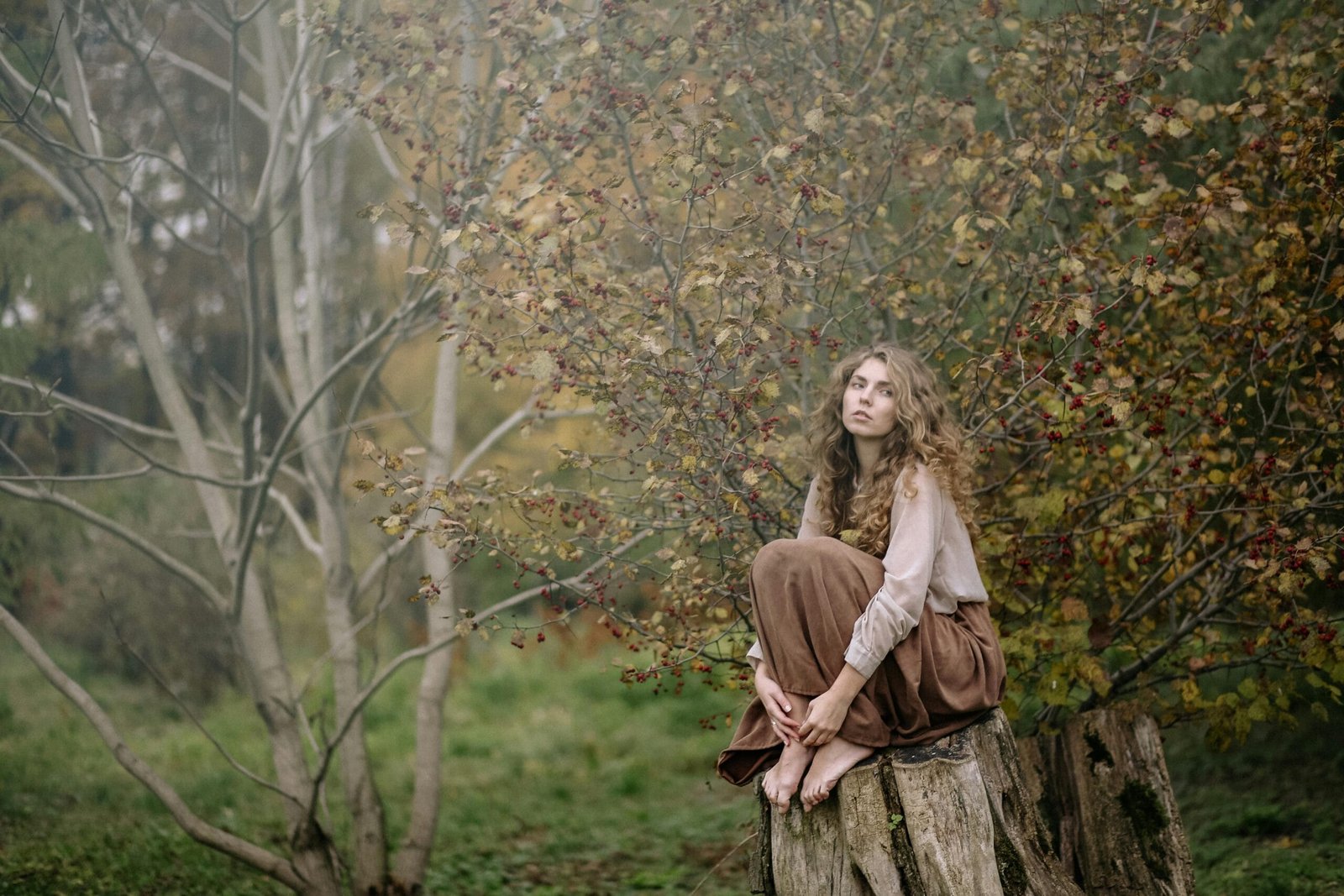 A thoughtful woman with curly hair sitting on a tree stump in an autumnal forest.