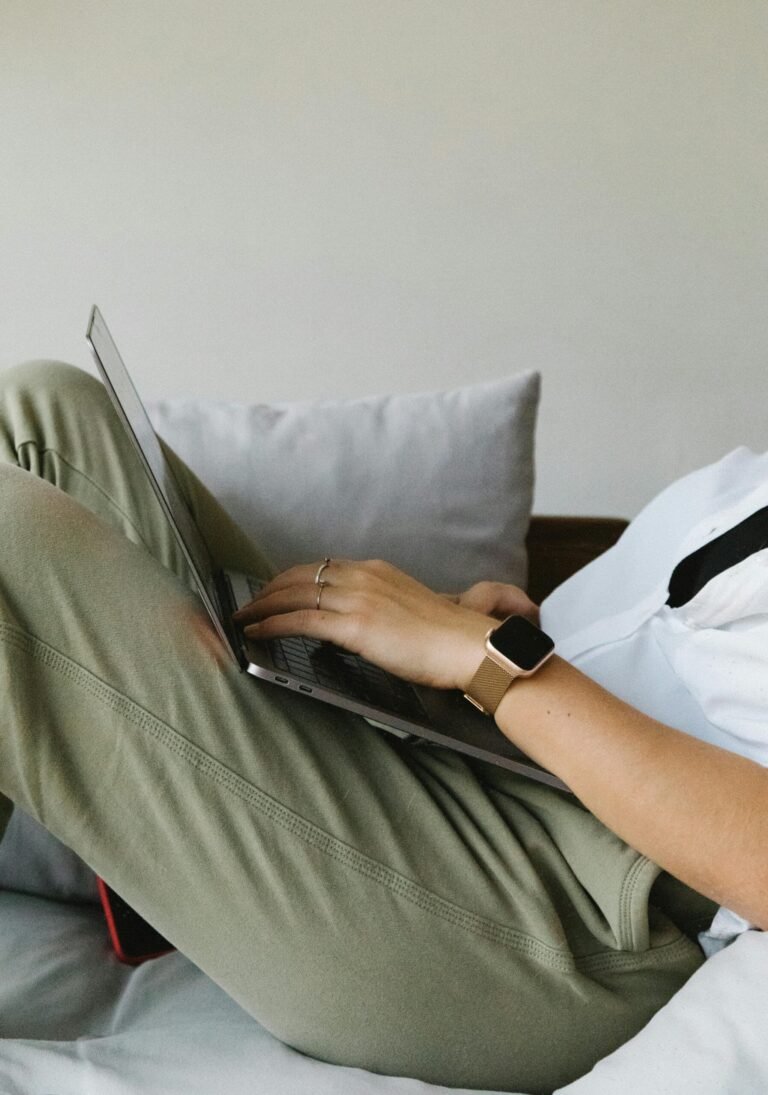 Side view of crop unrecognizable female freelancer in casual clothes and smartwatch lying on comfortable sofa and working remotely on laptop