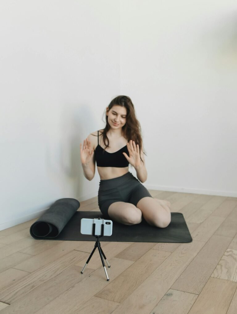 Woman on yoga mat recording a fitness video with her phone on a tripod at home.