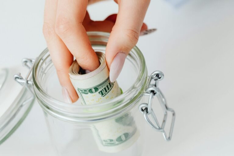 A close-up of a hand placing rolled dollars into a glass jar, symbolizing savings.